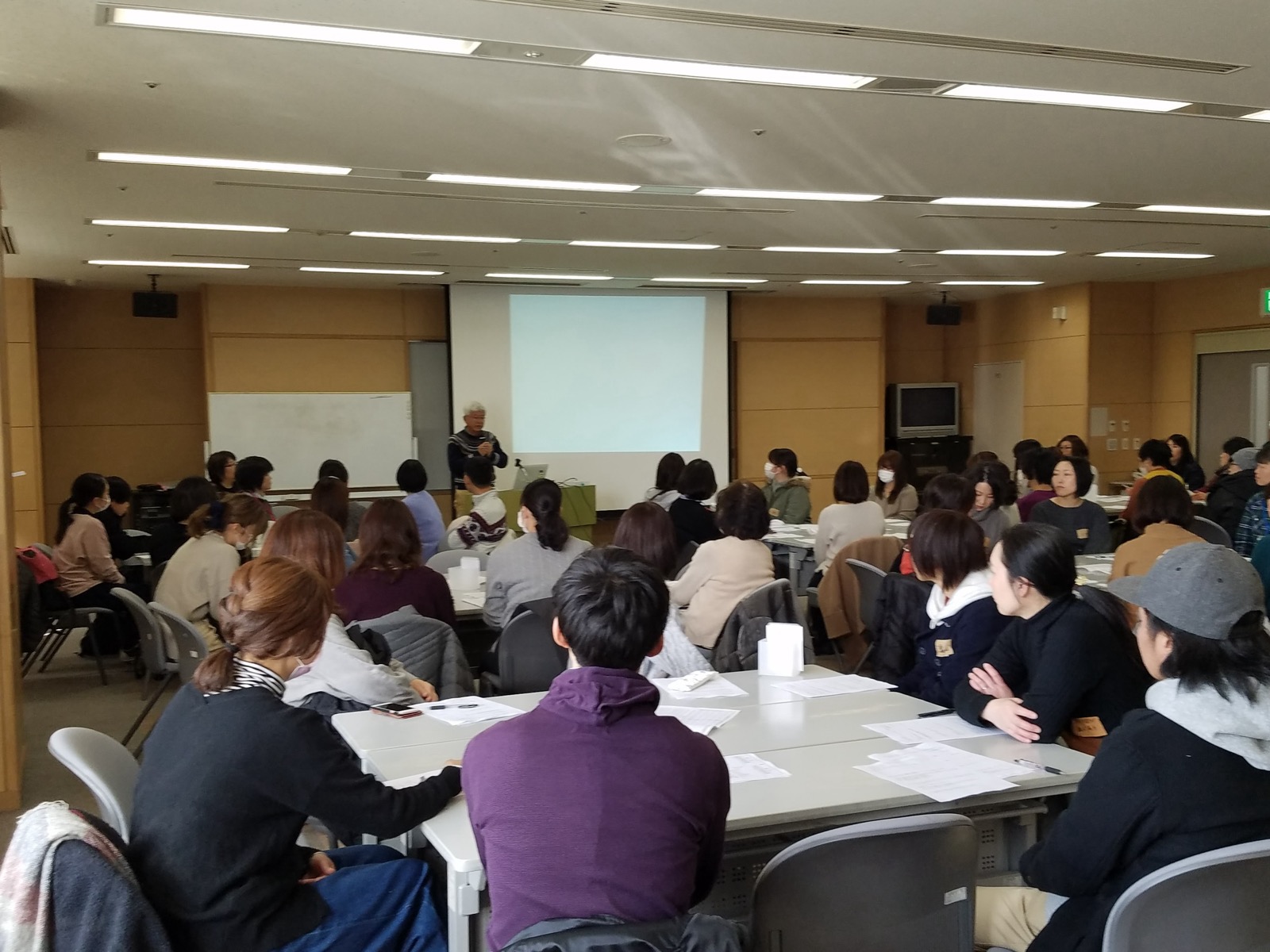 研修会場。前のスクリーンの横で講師が話し、机に着いた大勢の支援員が聞いている様子