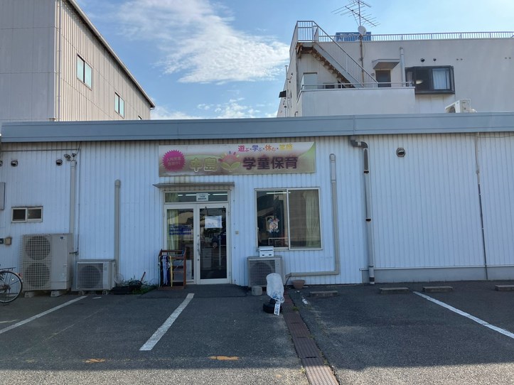白い波板の建物の入口を正面から見たところ。「中島学童保育 遊ぶ×学ぶ×休む×笑顔」の看板の下にガラスの引き戸があり、わきに傘立てと何本かのかさが置かれている