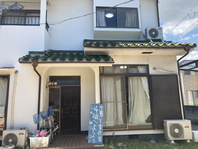 民家の玄関まわり。濃い茶色のドアの上に緑の瓦のひさし、右にレースのカーテンの掃き出し窓。玄関の右に「中島学童保育 4組へようこそ」と書かれた黒板。左手前に虫取り網とかご、おもちゃ箱、ボール