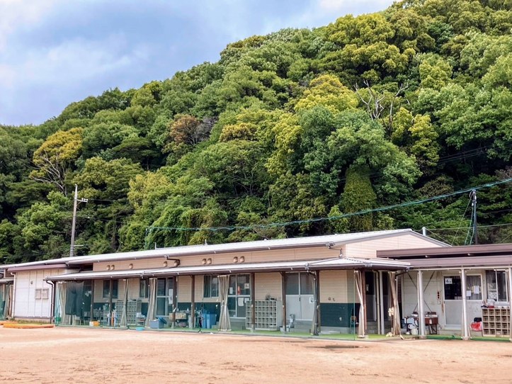 グラウンドの向こうに立つ、横に長い平屋のプレハブ2棟。軒下の通路に下駄箱や道具がならび、うしろに緑の木がしげる山がせまっている