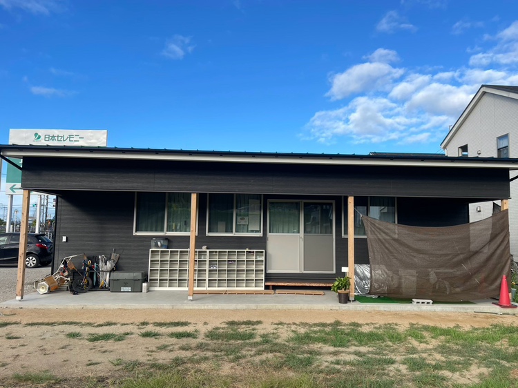 黒い板壁の平屋の建物。軒下に下駄箱がならび、右手に日よけの茶色い幕が張られている