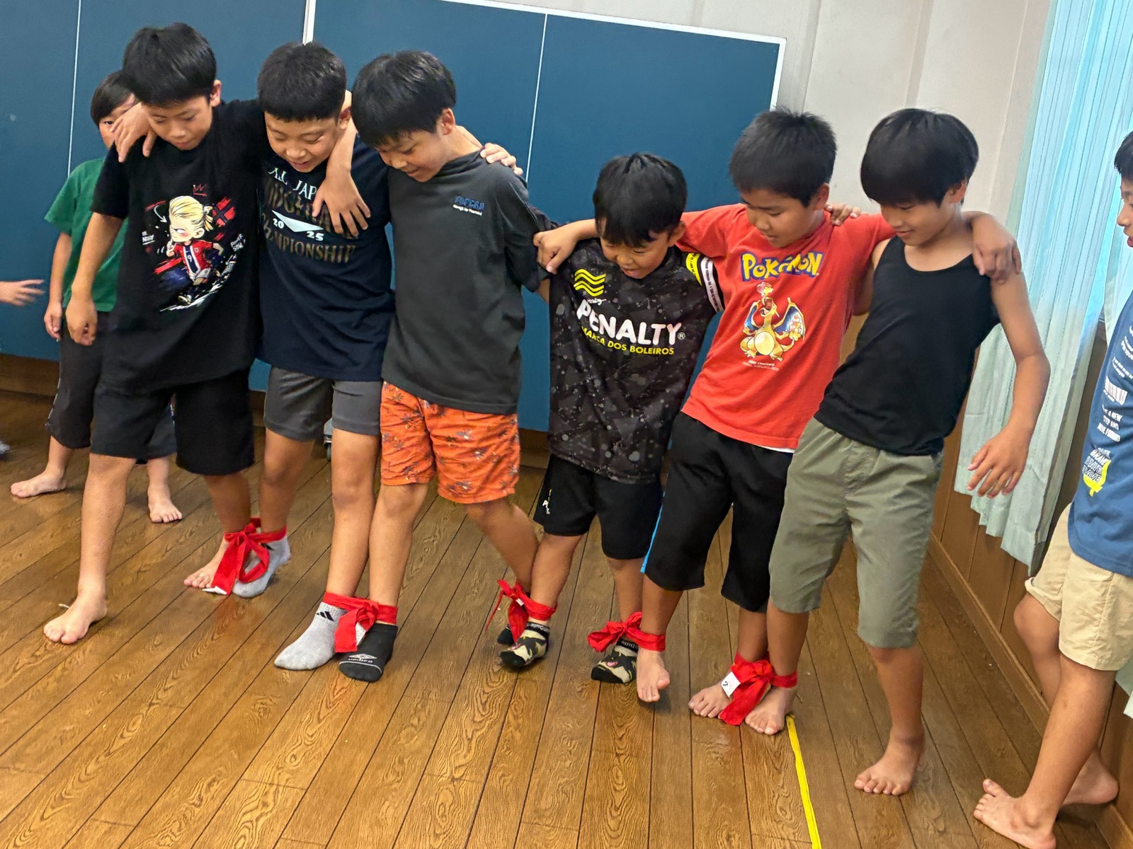 こどもたちが足首を赤いひもで結び、肩を組んで並んで歩こうとしている様子