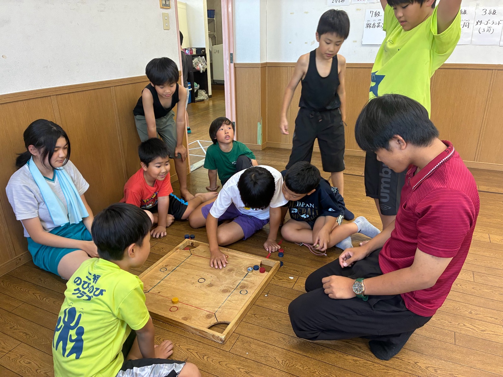 床に置いた木の盤でカロムをするこどもたちを、支援員とまわりのこどもがのぞきこんでいる様子