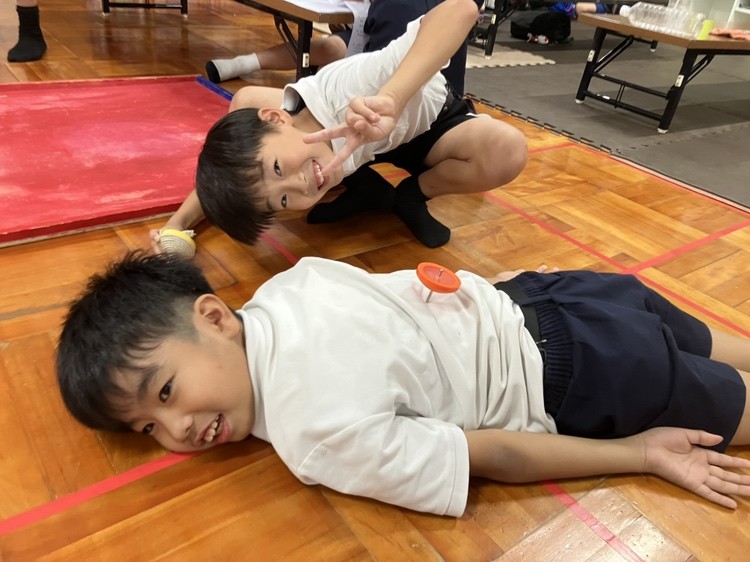 こどもがうつぶせになり、背中にのせたこまを回している様子